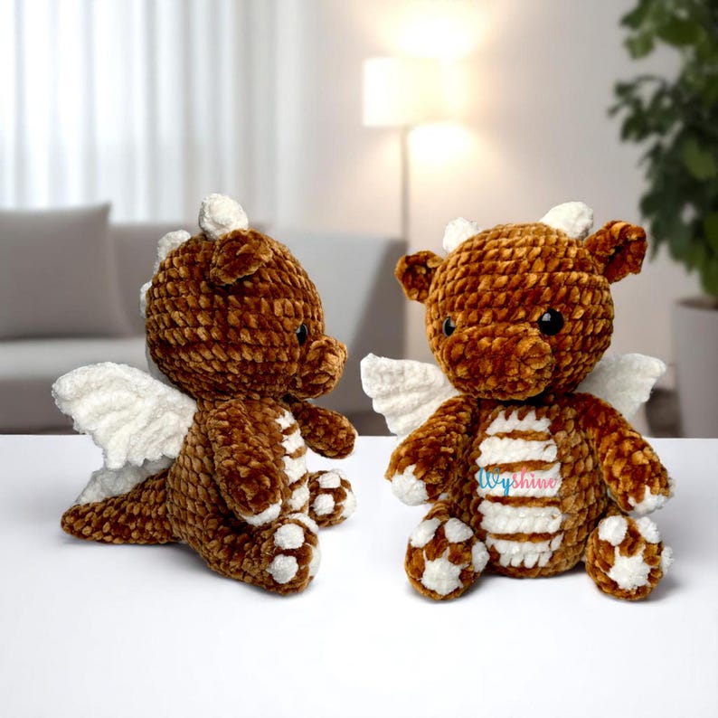 May include: Two handmade brown crochet dragon plushies with white wings and accents. The dragons have small black eyes and white horns. One dragon is lying down, and the other is sitting upright. The dragons are on a white surface.
