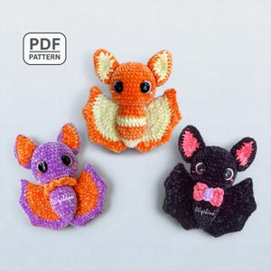 May include: Three handmade crochet bats in orange, purple, and black. The bats have large eyes and detailed wings. The orange bat has cream-colored wings, the purple bat has orange wings, and the black bat has pink accents. The image includes the text "PDF PATTERN".