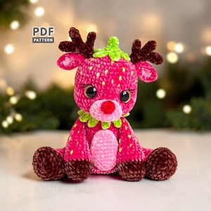 Puede incluir: Un peluche de ganchillo con forma de reno y diseño inspirado en una fresa. El cuerpo es rosa brillante con motas amarillas, vientre rosa claro, astas y pies marrón oscuro, nariz roja y cuello de hojas verdes. El texto dice PDF PATTERN.