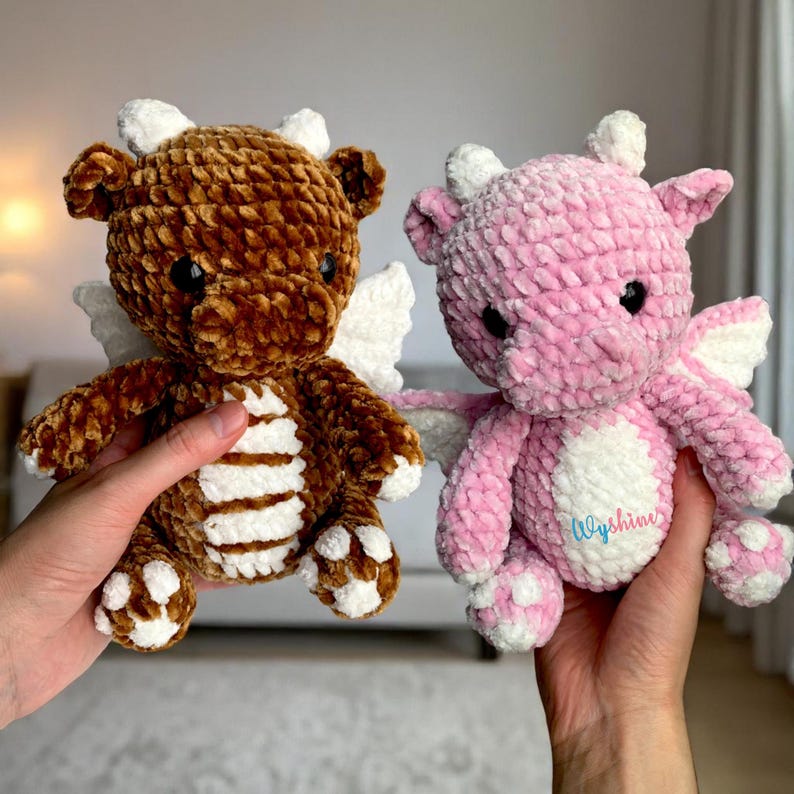May include: Two plush crochet dragons, one brown and one pink, are held up. The dragons have white wings, horns, and belly details. The pink dragon has the word "Wyshine" embroidered on its belly.