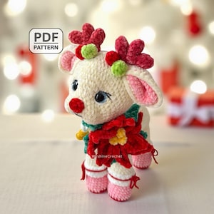 May include: A crocheted white reindeer with red and green details, wearing a red poinsettia and pink shoes. The reindeer has a red nose, blue eyes, and a green scarf. The text "PDF PATTERN" is in the upper left corner.