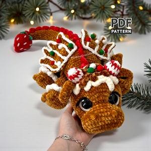 Könnte beinhalten: Ein gehäkelter Lebkuchen-Drache mit rotem Erdbeerschwanz, weißen Zuckerguss-Akzenten und dekorativen Süßigkeiten. Der Drache hat große schwarze Augen und ist mit "PDF PATTERN" beschriftet. Der Drache ist eine festliche Weihnachtsdekoration.