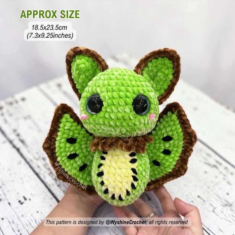 May include: A plush crochet bat toy featuring a bright green body, large black eyes, and brown-trimmed wings. The belly has a yellow and black kiwi fruit pattern. It measures approximately 7.3 by 9.25 inches. Text on image: APPROX SIZE 18.5x23.5cm (7.3x9.25inches).