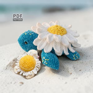 May include: A crocheted turtle with a large white and yellow daisy flower on its back and a smaller daisy next to it. The turtle is blue with yellow accents. The text "PDF PATTERN" is in the upper left corner.
