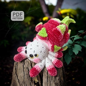May include: A handmade crocheted dragon fruit spider toy. The spider has a white body, pink and red legs, and a dragon fruit head. The toy is on a tree stump. The image includes a "PDF PATTERN" logo.