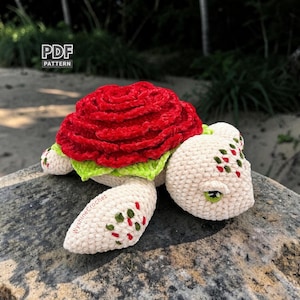 Puede incluir: Una tortuga de ganchillo hecha a mano con un caparazón en forma de rosa roja, ribete verde y cuerpo blanco roto. La cabeza y las extremidades están decoradas con detalles rojos y verdes. Incluye el texto "PDF PATTERN".
