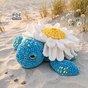 May include: A plush, blue turtle with a large white and yellow daisy on its back. The turtle's shell and flippers have yellow accents. The text "PDF PATTERN" is in the upper left corner. The turtle is on a sandy surface.