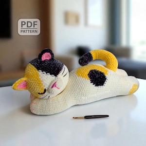 Puede incluir: Un juguete de gato tejido a ganchillo en crema, amarillo, negro y rosa. El gato está acostado con los ojos cerrados, y hay un ganchillo al lado. La cabeza del gato es negra y amarilla, con orejas rosas. La imagen incluye el texto "PDF PATTERN".