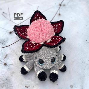 May include: A crochet plush toy featuring a gray spider-like body with black and white tipped legs, large blue eyes, and a top section resembling a dark red flower with white spots and a textured, light pink brain-like center. Text reads PDF PATTERN.