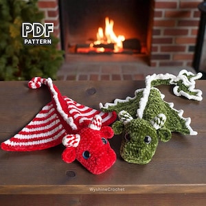 May include: Two crocheted dragon toys. One is red with white stripes and accents, the other is green with white accents. The dragons have large eyes and are displayed on a wooden surface. A fireplace with a burning fire is in the background.