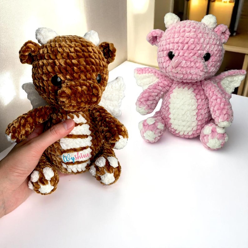 May include: Two plush dragon toys. One is brown with white accents and wings, the other is pink with white accents and wings. The brown dragon is being held by a hand. The dragons have small horns and are sitting on a white surface.