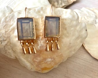 Könnte beinhalten: Ein Paar goldfarbene, rechteckige Ohrhänger. Jeder Ohrring hat einen hellfarbigen, rechteckigen Stein mit geometrischem Design und drei kleine, baumelnde, glockenförmige Anhänger. Die Ohrringe werden auf einer Muschel präsentiert.