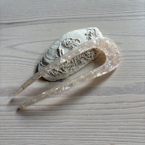 May include: A pale pink hair fork with a marbled design, placed on a textured, off-white stone. The hair accessory is curved with two long prongs. The text "CREATION FRANCE" is visible on the hair fork.