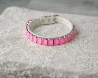 Puede incluir: Un collar de perro de cuero blanco con piedras de imitación cuadradas de color rosa.
