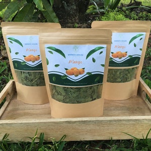 May include: Three brown paper pouches filled with green leaves, labeled "Mango" with a mango illustration. The pouches sit on a wooden tray, with a clear window showing the contents. The background is green grass and a tree.