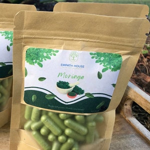 Op de afbeelding: Een bruine papieren zak met een doorzichtig plastic venster, met groene capsules erin. De zak heeft een label met de tekst "Empath House" en "Moringa". Het label vermeldt ook "Gekweekt in de achtertuin, gerijpt in de zon van Florida!"