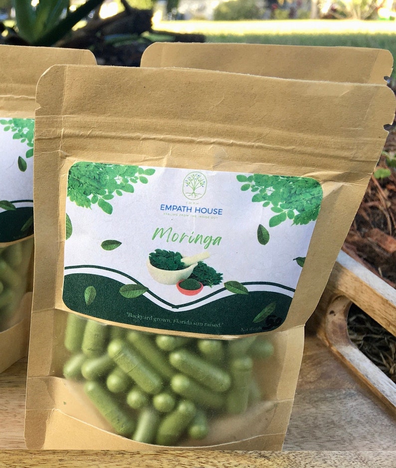 Op de afbeelding: Een bruine papieren zak met een doorzichtig plastic venster, met groene capsules erin. De zak heeft een label met de tekst "Empath House" en "Moringa". Het label vermeldt ook "Gekweekt in de achtertuin, gerijpt in de zon van Florida!"