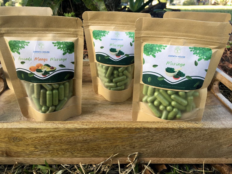 Op de afbeelding: Drie bruine papieren zakken gevuld met groene capsules. De zakken hebben etiketten met de tekst "Empath House" en verschillende productnamen: "Avocado Mango Moringa", "Moringa" en "Moringa".