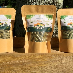 Puede incluir: Tres bolsas de papel marrón con ventanas transparentes, cada una llena de cápsulas verdes. Las bolsas tienen etiquetas con el texto "EMPATH HOUSE Avocado Mango Moringa" y un gráfico de aguacates y mangos. Las bolsas están sobre una superficie de madera.