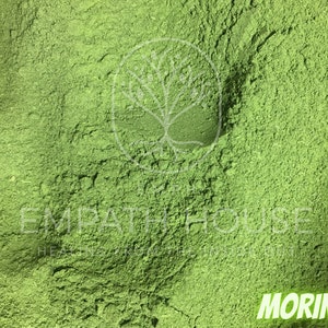 Op de afbeelding: Close-up van een felgroen poeder met het label "MORINGA". Het poeder is fijn gemalen en heeft een licht getextureerd oppervlak.