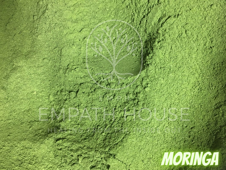 Op de afbeelding: Close-up van een felgroen poeder met het label "MORINGA". Het poeder is fijn gemalen en heeft een licht getextureerd oppervlak.