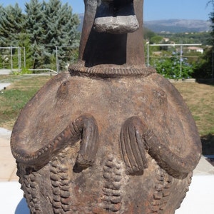 Puede incluir: Una escultura de terracota de una figura humana estilizada con una cabeza grande y una boca ancha. La figura lleva una prenda decorativa y tiene los brazos cruzados sobre el pecho. La escultura es de color marrón y tiene una textura rugosa.