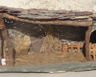May include: A miniature nativity scene made of natural materials, including wood, stone, and moss. The scene features a small wooden stable with a thatched roof, a wooden fence, and a small white lamb.