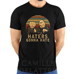 May include: Black t-shirt with a retro design. The design features two cartoon characters and the text "HATERS GONNA HATE" in gold. The background has orange, brown, and yellow stripes, resembling a sunset.