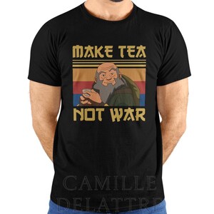 Pode incluir: Uma t-shirt preta de manga curta com uma estampa gráfica de um homem idoso com barba segurando uma xícara de chá. O design inclui listras horizontais em amarelo, laranja e azul, com o texto "MAKE TEA" acima e "NOT WAR" abaixo da imagem.