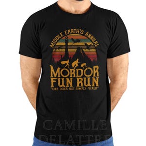 Puede incluir: Camiseta negra con un diseño gráfico de estilo vintage. El diseño presenta el texto "Middle Earth's Annual Mordor Fun Run" con una montaña y siluetas. También incluye la frase "One Does Not Simply Walk".
