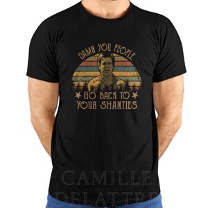 Puede incluir: Camiseta negra con un diseño retro que presenta la cara de un hombre dentro de un gráfico de puesta de sol. El texto dice "Damn You People Go Back To Your Shanties". Una divertida camiseta gráfica de estilo vintage.