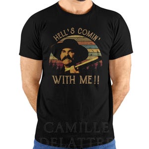 May include: Black t-shirt featuring a vintage-style graphic of a man in a cowboy hat with a mustache, holding a gun. The text "HELL'S COMIN' WITH ME!!" is above and below the image. The shirt is a classic crew neck.