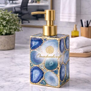Puede incluir: Dispensador de jabón cuadrado con bomba dorada, adornado con rodajas de ágata azul y turquesa, con bordes dorados. El texto "Omsaihandicraft" es visible. Un elegante accesorio de baño.