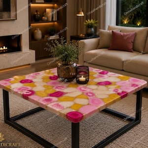 May include: A square coffee table with a colourful agate stone top in shades of pink, yellow, and white. The table has a black metal frame and is in a living room setting. A decorative vase and candle are on the table.