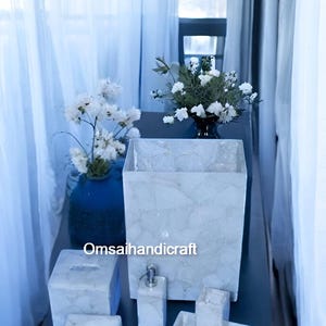 May include: A set of white marble bathroom accessories, including a soap dispenser, a toothbrush holder, a soap dish, and a tissue box. The accessories are arranged on a dark blue surface. The text "Omsaihandicraft" is visible in the image.