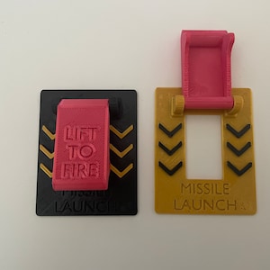 May include: Two 3D-printed missile launch control panels. One is black with a pink "Lift to Fire" lever and gold arrows. The other is gold with a pink lever and the text "Missile Launch".
