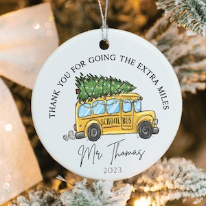 Puede incluir: Adorno de cerámica blanca con un autobús escolar amarillo que lleva un árbol de Navidad. El texto dice "Gracias por hacer el esfuerzo extra" y "Mr. Thomas 2023".