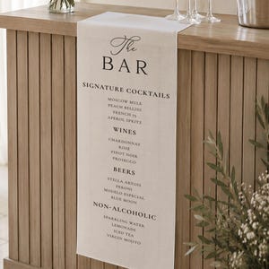 Può includere: Un runner da bar in tessuto bianco drappeggiato su un bancone in legno, con testo stampato in nero che recita The Bar, Signature Cocktails, Wines, Beers e Non-Alcoholic, seguito da un elenco di varie opzioni di bevande in un carattere serif pulito.