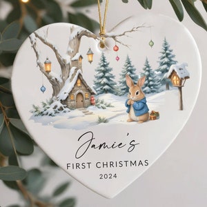 May include: A white heart-shaped ceramic bauble with a winter scene featuring a small cottage, a rabbit in a blue coat, and snow-covered trees. The bauble has the text "Jamie's First Christmas 2024" written on it.