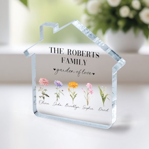Könnte beinhalten: Ein klares, acryl-geformtes Haus-Ornament mit dem Text "THE ROBERTS FAMILY garden of love" und fünf bunten Blumen mit Namen darunter: Olivia, John, Brooklyn, Sophia, David.