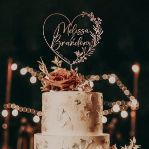 May include: A pink heart-shaped cake topper with the text "Melissa & Brandon" and the date "05.05.2024". The topper is decorated with floral details and is placed on top of a white wedding cake.
