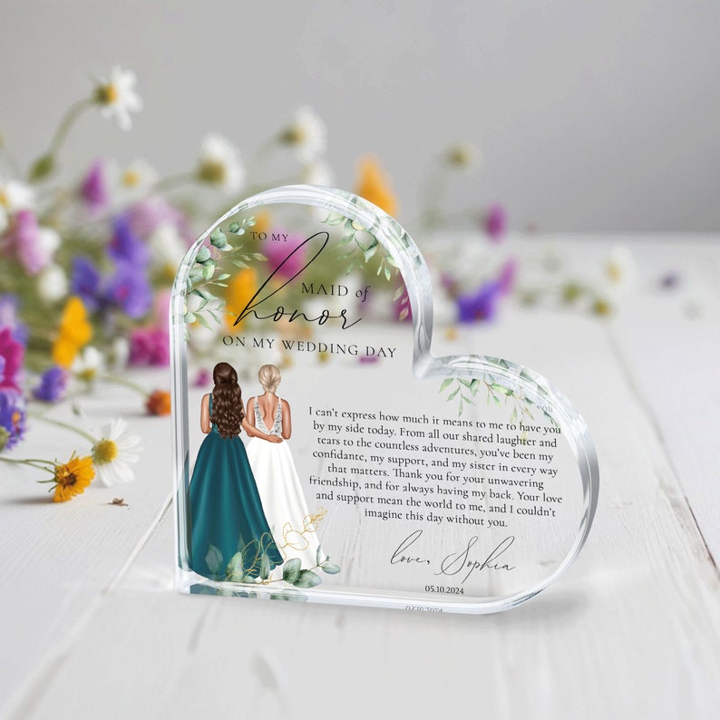 May include: A heart-shaped acrylic keepsake with a floral design and two women in wedding attire. The text reads "To my Maid of Honor on my Wedding Day". The text continues with a message of appreciation for the maid of honor and is signed "Love, Sophia" with a date.