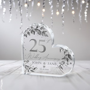 May include: A clear heart-shaped crystal paperweight with a silver design. The paperweight features the text "25th Wedding Anniversary John & Jane 09.18.1999".