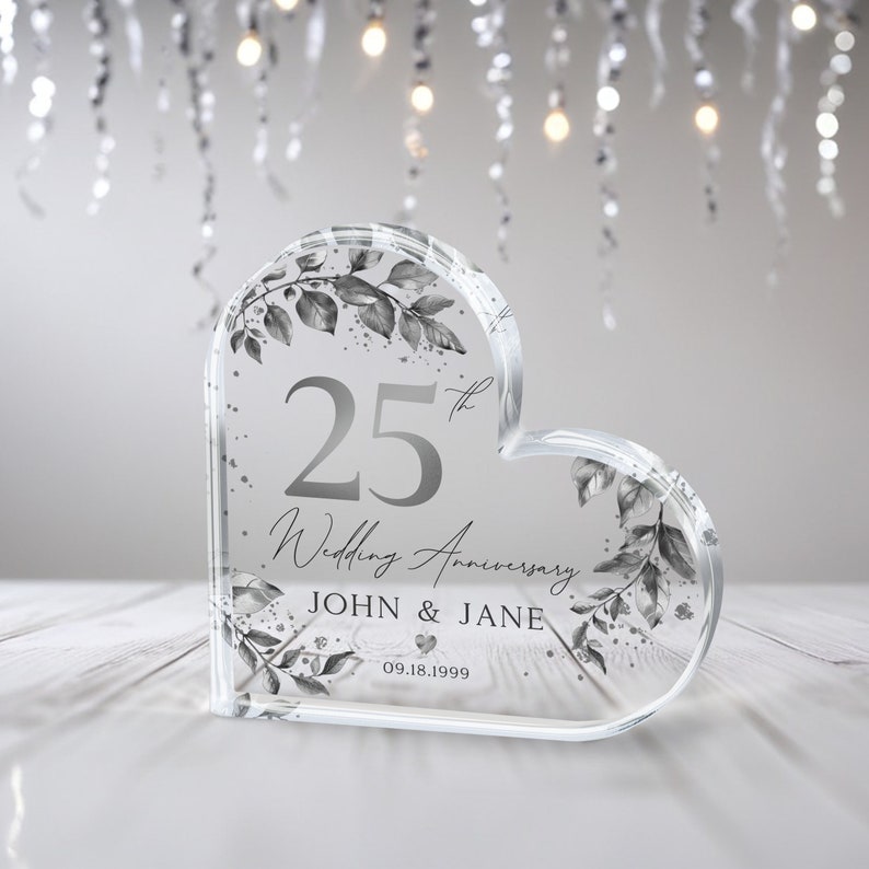May include: A clear heart-shaped crystal paperweight with a silver design. The paperweight features the text "25th Wedding Anniversary John & Jane 09.18.1999".
