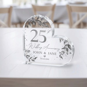 May include: A clear heart-shaped crystal paperweight with a silver design. The paperweight features a floral pattern, the number 25, the text "Wedding Anniversary", the names "JOHN & JANE", and the date "09.18.1999".