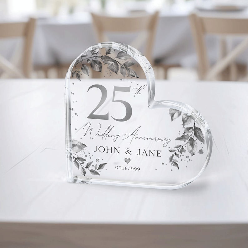 May include: A clear heart-shaped crystal paperweight with a silver design. The paperweight features a floral pattern, the number 25, the text "Wedding Anniversary", the names "JOHN & JANE", and the date "09.18.1999".