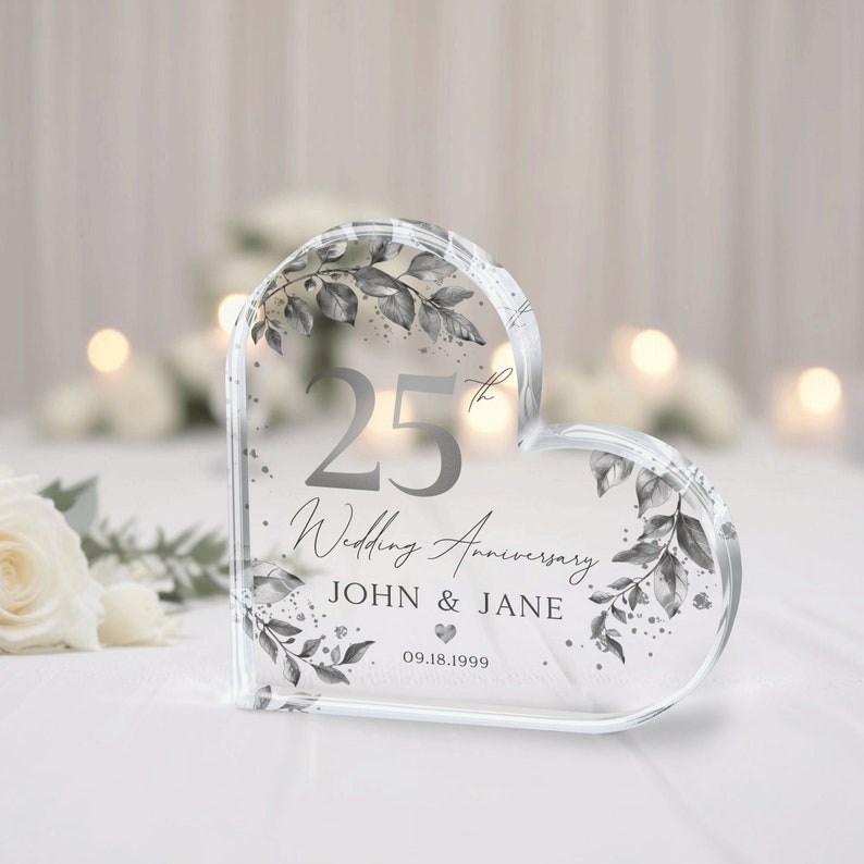 May include: A clear heart-shaped crystal paperweight with a silver design. The paperweight features the number 25, the words "Wedding Anniversary", the names "JOHN & JANE", and the date "09.18.1999".