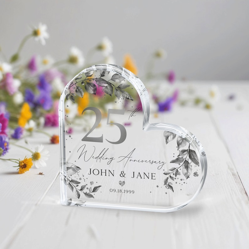 May include: A clear heart-shaped acrylic keepsake with a silver design. The design features a floral pattern, the number 25, the words "Wedding Anniversary", and the names "JOHN & JANE" with the date "09.18.1999".