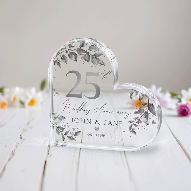 May include: A clear heart-shaped acrylic paperweight with a silver design. The paperweight features a floral design with the number 25, the text "Wedding Anniversary", the names "JOHN & JANE", and the date "09.18.1999".