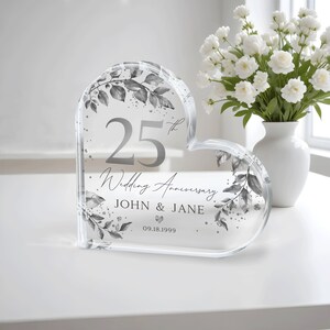 May include: A clear heart-shaped crystal paperweight with a silver design of leaves and the text "25th Wedding Anniversary John & Jane 09.18.1999".